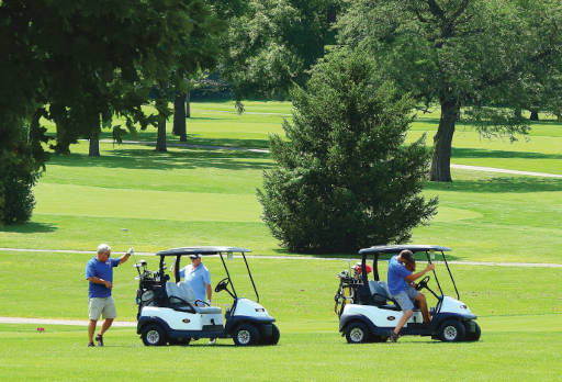 Two golf carts in the foreground on a golf course. One person is entering the golf cart on the right. A second person is selecting a club from the bags on the left-hand cart. A third person stands behind the left cart.